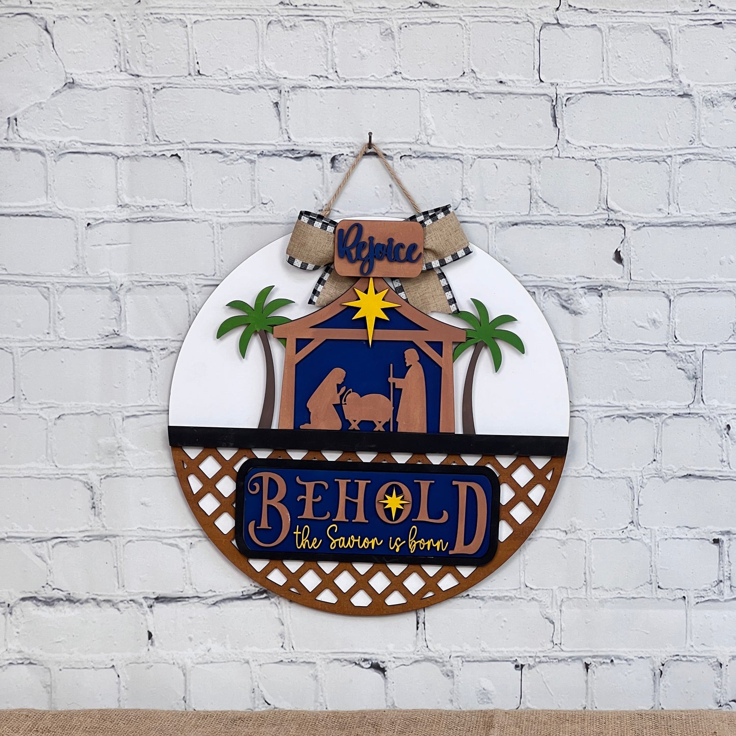 The DIY Door Hanger Interchangeable Base by Janet's Craft Corner features a nativity scene on a round insert with "Rejoice" on a bow, "Behold the Savior is born," and two palm trees. Perfect for festive charm against a white brick wall or door.