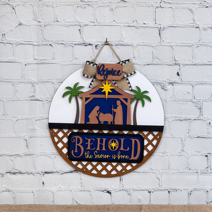 The DIY Door Hanger Interchangeable Base by Janet's Craft Corner features a nativity scene on a round insert with "Rejoice" on a bow, "Behold the Savior is born," and two palm trees. Perfect for festive charm against a white brick wall or door.