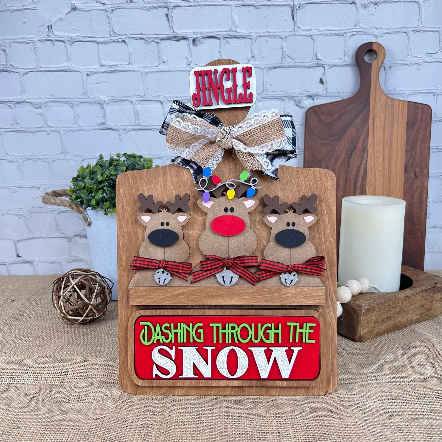 Janet's Craft Corner presents the Breadboard Interchangeable Base with Insert. This DIY home decor kit includes a wooden holiday piece featuring three reindeer with plaid bows, a "Jingle" sign, and "Dashing Through the Snow," complemented by accessories like a candle, plant, and decorative ball.