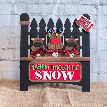 Janet's Craft Corner offers a Garden Fence with Insert DIY kit featuring three cartoon reindeer with red noses and plaid scarves. It includes "Jingle" and "Dashing Through the Snow" signs, interchangeable inserts, and colorful lights for festive charm.