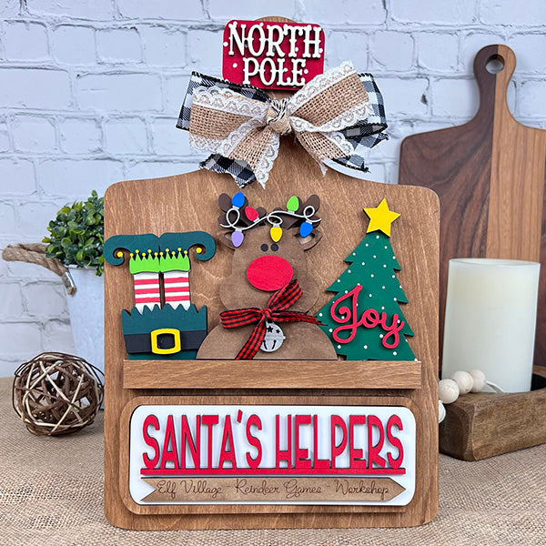 Create festive DIY decor with Janet's Craft Corner's Breadboard Interchangeable Base, featuring inserts like a reindeer with Christmas light antlers, elf hat and shoes, "Joy" tree, "SANTA'S HELPERS" text in red, topped with a ribbon-adorned "NORTH POLE" plaque.