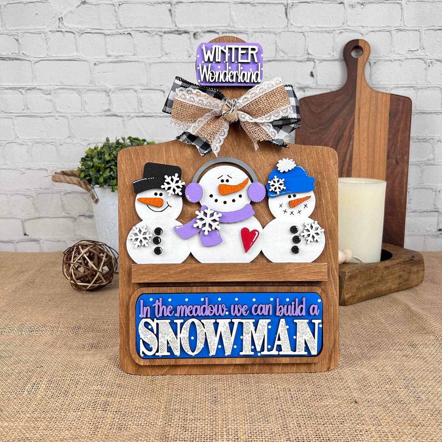 Janet's Craft Corner offers a Breadboard Interchangeable Base with Insert DIY kit. The base features three snowmen: one in a black hat, one in a purple scarf, and one in a blue hat. Signs read "Winter Wonderland" and "In the meadow we can build a snowman," making it perfect for festive home decor.