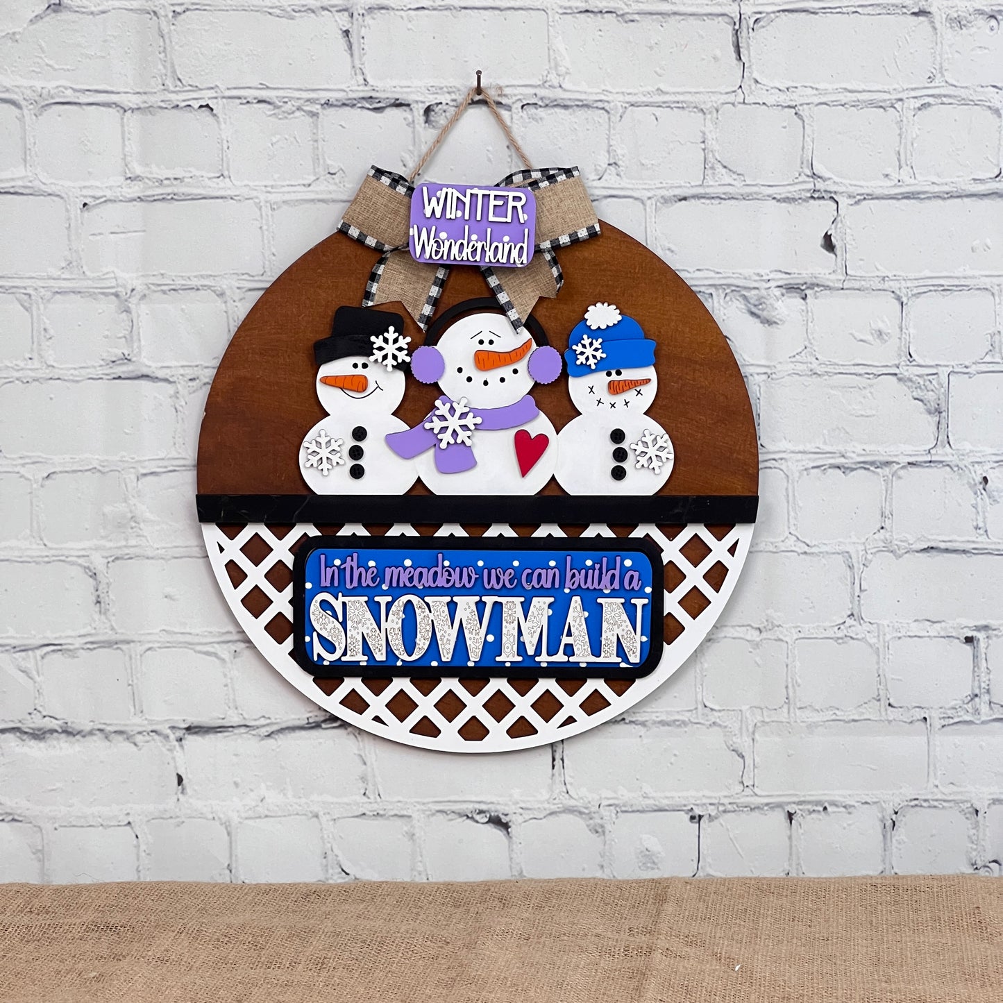 The DIY decorative wall hanging from Janet's Craft Corner features three accessorized snowmen, ideal for home décor. The top reads "Winter Wonderland," and the bottom says "In the meadow, we can build a snowman" on a blue sign. This interchangeable door hanger enhances any white brick wall.