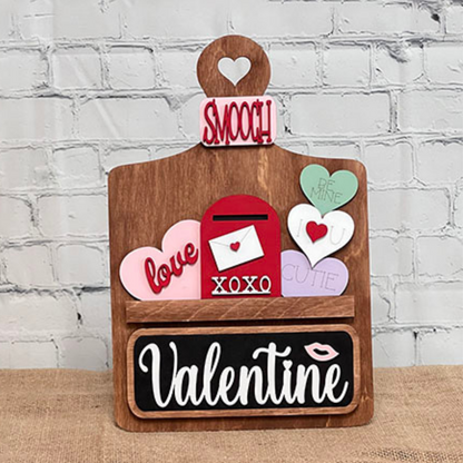 Janet's Craft Corner offers a DIY home decor kit called the Breadboard Interchangeable Base with Insert. It includes a wooden base shaped like a cutting board, featuring "Smooch" on top, interchangeable accessories, and designs like hearts with words like "Be Mine," plus "XOXO" and "Valentine.