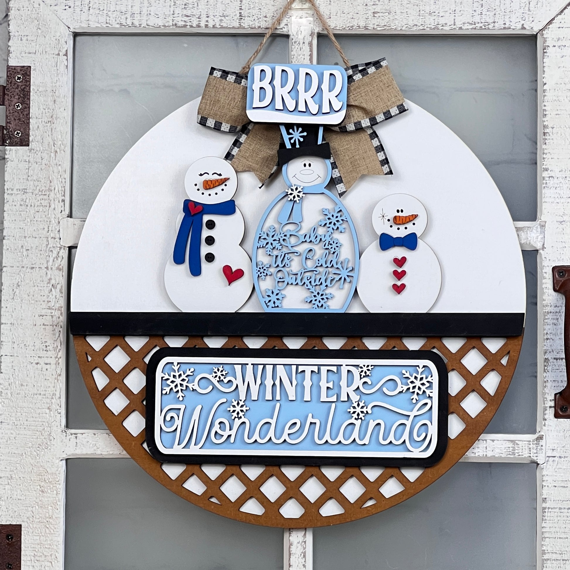 Janet's Craft Corner offers a Door Hanger Interchangeable Base with Insert, a DIY kit featuring winter charm. The design showcases three snowmen, "BRRR" and "Let it Snow," while "Winter Wonderland" appears below with snowflakes in a white, blue, and brown palette accented by decorative ribbons.