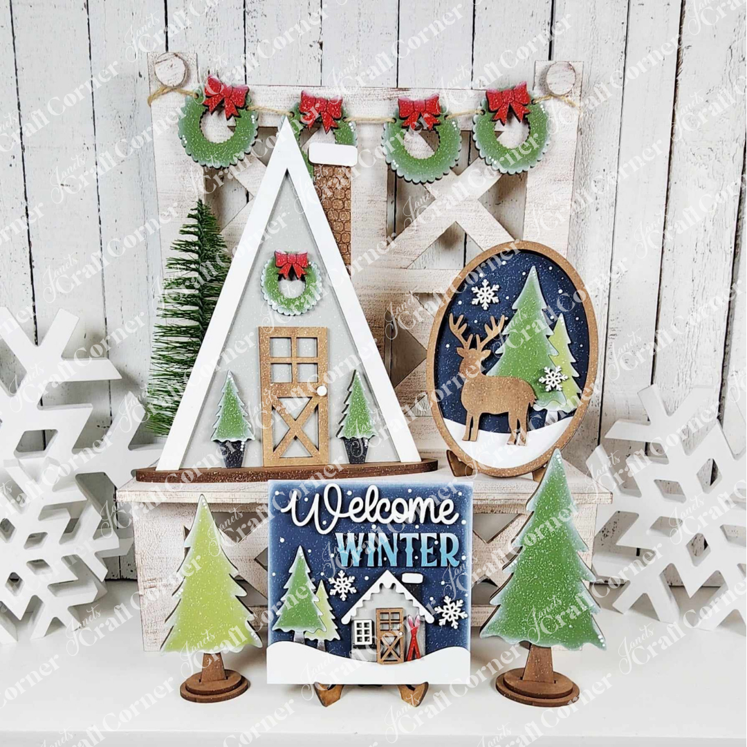 A cozy winter-themed display showcasing a hand-painted, house-shaped decor piece, a charming "Welcome Winter" sign, and an oval design featuring deer and snowflakes is part of the Winter Cabin Decor Set from Janet's Craft Corner. This DIY craft kit also includes small green trees and snowflake decorations set against a rustic wood background.