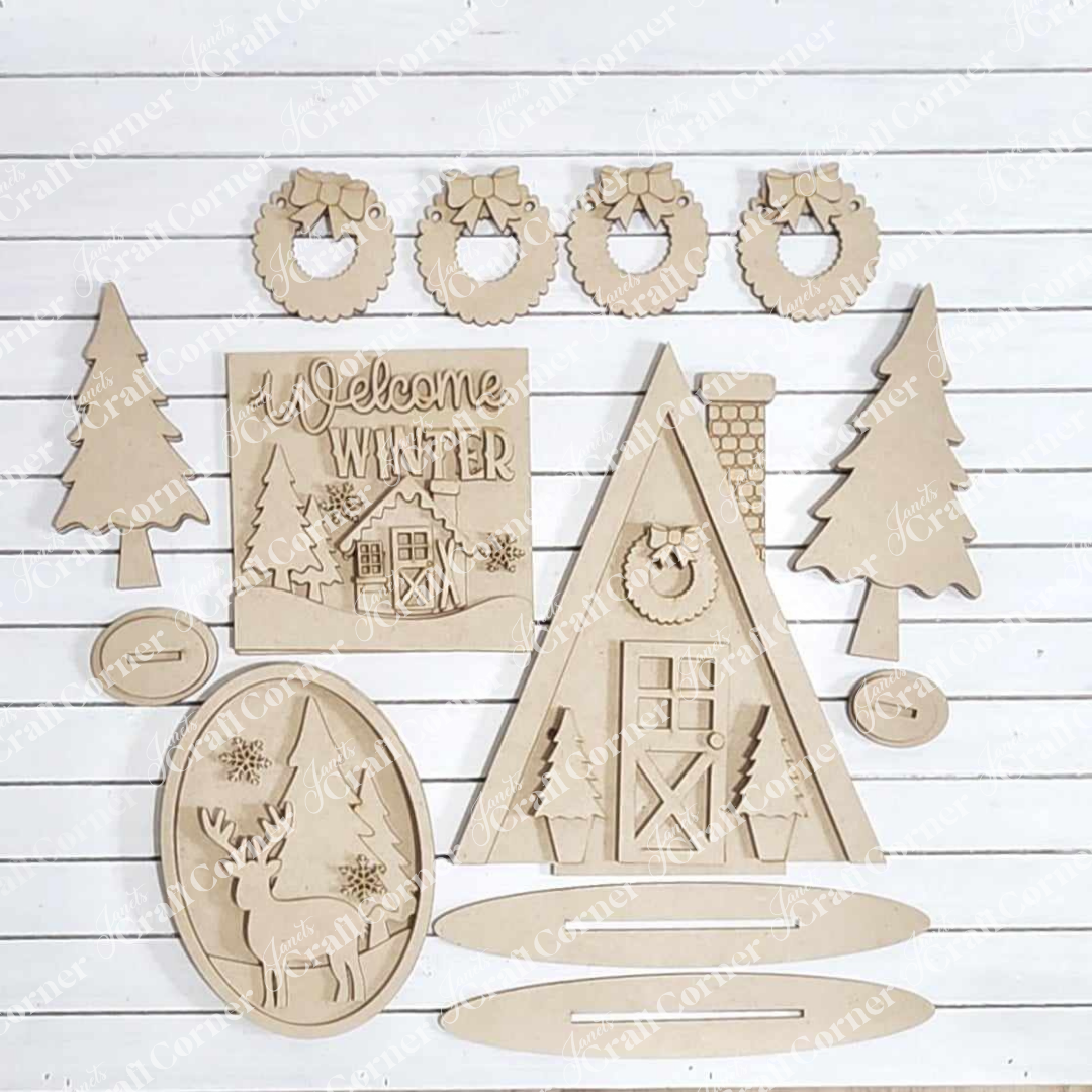 The Winter Cabin Decor Set from Janet's Craft Corner showcases wooden cutouts on a white background, featuring a winter-themed house, pine trees, wreaths, and a plaque with "Welcome Winter" text. This DIY craft kit includes hand-painted deer and geometric shapes for assembling stunning home décor.