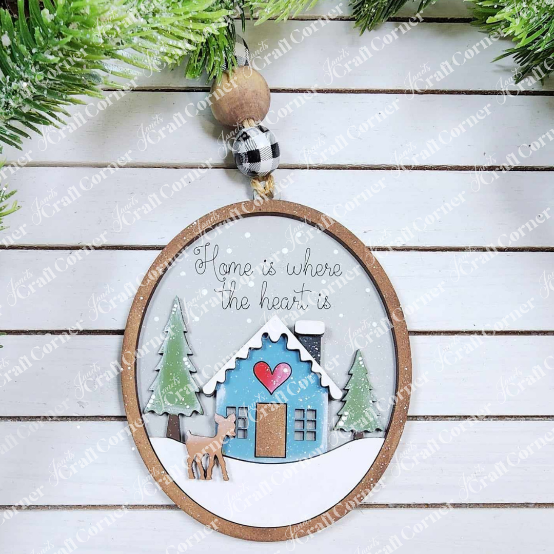 This delightful Winter Cabin Ornament, from Janet's Craft Corner, includes a charming design of a blue house, two green trees, and a brown deer in snow. Enhanced with a red heart and the phrase "Home is where the heart is," it gracefully hangs against a wooden backdrop with greenery.