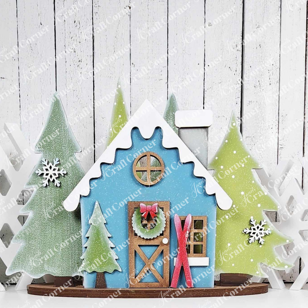 A charming festive scene, reminiscent of Janet's Craft Corner's Winter Cabin Votive Holder, showcases a miniature blue house with a snowy roof. This handmade home decor piece includes delightful touches like a wreath on the door and red skis leaning against it. Snowflakes complete the enchanting setup, surrounded by snow-covered trees and set against a white wooden background.