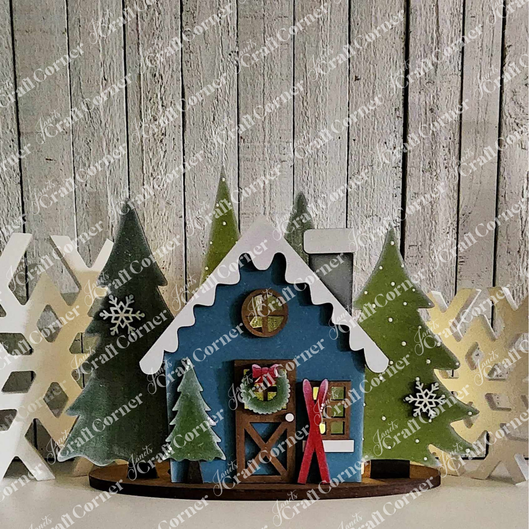 A decorative winter scene features a blue wooden house with the charm of Janet's Craft Corner's Winter Cabin Votive Holder, complete with skis and snowflakes, nestled among pine trees. The snow-topped roofs and large cutouts enhance the festive allure of this handmade piece against a rustic wooden plank backdrop.