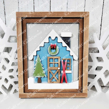 Embrace the charm of winter with Janet's Craft Corner's Winter Cabin Interchangeable Farmhouse Sign DIY Home Decor Craft Kit. This set features a blue house with a snow-covered roof and chimney, complemented by a hand-painted wooden door adorned with a welcoming green wreath. Skis rest beside it against a crisp white backdrop, complete with an evergreen pine tree.