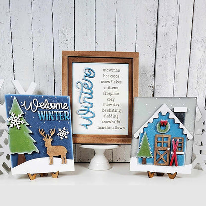 Displayed against a white rustic wooden backdrop are three winter-themed decorations: Janet's Craft Corner's hand-painted "Welcome Winter" sign featuring a deer, a framed list of winter words, and a house adorned with snow and skis. These items make for perfect home decor pieces.