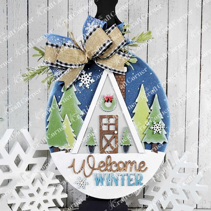 Introducing the Winter Cabin Wall Hanger from Janet's Craft Corner: This round winter-themed decoration showcases a hand-painted triangular house set among trees and snowflakes, adorned with a plaid and burlap bow. The kit features the words "Welcome Winter" at the bottom, and its wooden surface background, embellished with decorative snowflakes, adds personalized charm to your home décor.