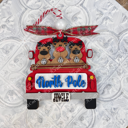A delightful ornament from Janet's Craft Corner, the Antique Truck Ornament is part of their DIY Decoration Kits. It showcases three reindeer in an antique red truck with a "North Pole" sign and a "Jingle" license plate, adorned with a red and green plaid bow on top against a textured white background. Perfect for adding charm to your Christmas décor collection.