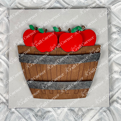 Janet's Craft Corner presents Apple Basket Square: a wood cut-outs craft kit featuring a wooden basket filled with bright red apples and green leaves, ideal for seasonal DIY decor, displayed on a textured white background.