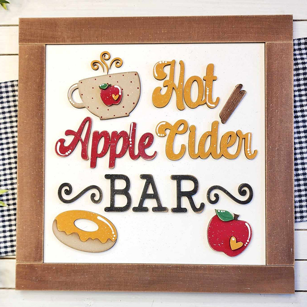 The Apple Cider Bar Sign Shelf Sitter from Janet's Craft Corner showcases decorative text alongside a steaming mug, cinnamon stick, donut, and apple. Its wooden border and black-and-white checkered background make it a perfect addition for home décor enthusiasts who love DIY craft kits.