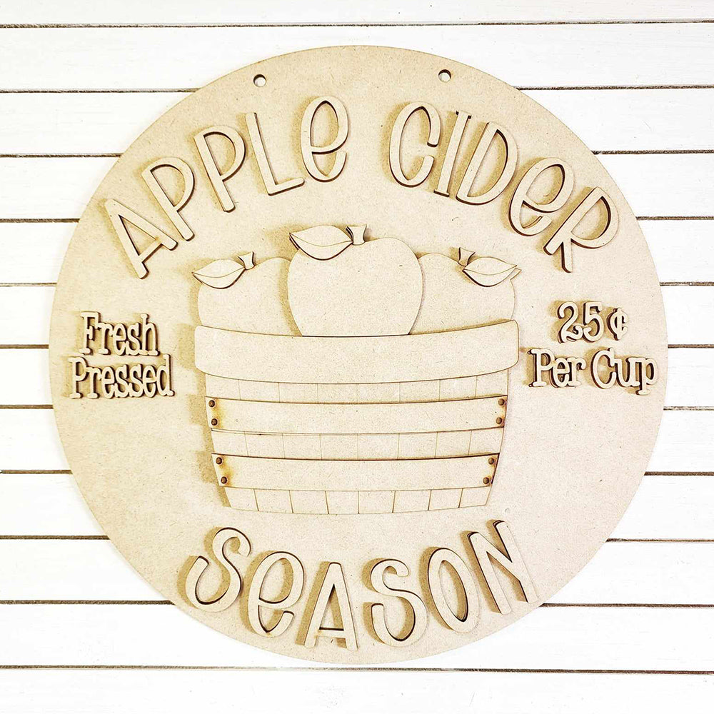 Introducing a delightful home décor piece from Janet's Craft Corner: the Apple Cider Door Hanger DIY home decor kit. This circular, wooden sign beautifully showcases "Apple Cider Season" alongside a basket of apples. Expertly hand-painted, it features the phrases "Fresh Pressed" and "25¢ Per Cup" on either side, making it an ideal addition to any space with its chic white striped background.