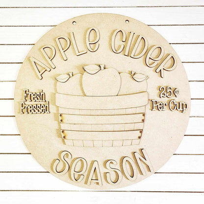 Introducing a delightful home décor piece from Janet's Craft Corner: the Apple Cider Door Hanger DIY home decor kit. This circular, wooden sign beautifully showcases "Apple Cider Season" alongside a basket of apples. Expertly hand-painted, it features the phrases "Fresh Pressed" and "25¢ Per Cup" on either side, making it an ideal addition to any space with its chic white striped background.