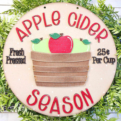 The Apple Cider Door Hanger from Janet's Craft Corner is a delightful DIY home decor kit that features a round sign with green leaves and the phrase "Apple Cider Season" in bold red text. Adorned with apples in a wooden basket, it also includes the messages "Fresh Pressed" and "25¢ Per Cup," adding an inviting charm to any space.