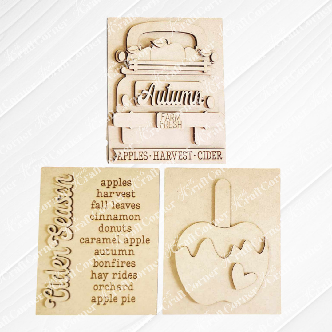 Displayed against a white plank wall this set of Apple Cider Switchable Farmhouse Signs are DIY seasonal home decor kits with a fall apple theme. One features the wood cut-out word “Cider Season” with words that evoke the scent of apples in the air, another features a caramel apple complete with heart and the last features an antique truck with a load of apples.