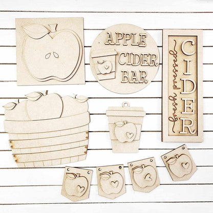 The Apple Cider Decor Set from Janet's Craft Corner features a charming collection of 6 wooden cutouts, each hand-painted with delightful apple-themed designs. This DIY home decor kit includes an apple shape, an "Apple Cider Bar" sign, a basket of apples, a coffee cup, and a banner adorned with apple icons. Perfect for craft enthusiasts looking to enhance their space with unique touches on a white striped background.