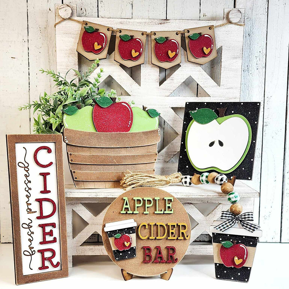 The Apple Cider Decor Set from Janet's Craft Corner, a DIY home decor kit featuring six pieces, includes a basket with red and green apples, a "Cider" sign, an apple-themed banner, and hand-painted apple ornaments. These charming elements are all beautifully arranged on a rustic white wooden background.