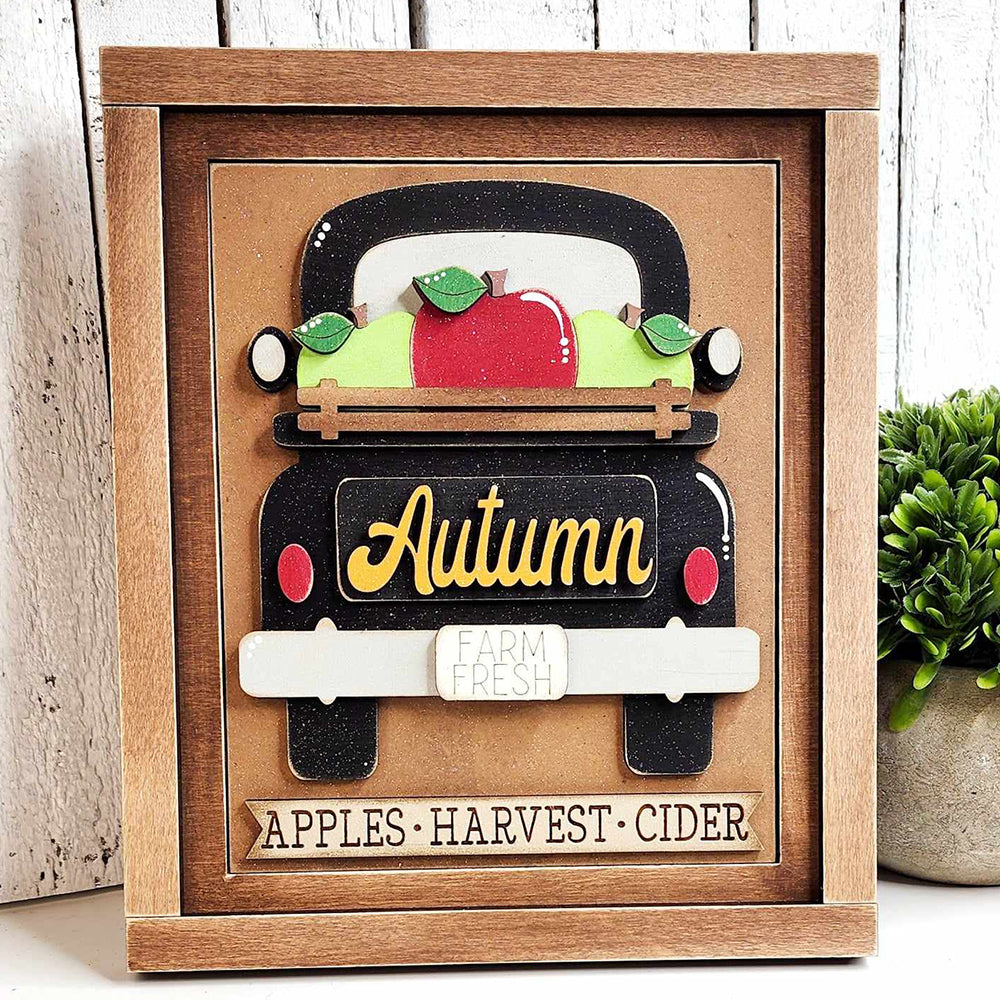 An Autumn Truck Interchangeable Sign from Janet's Craft Corner features a vintage truck overflowing with apples and pumpkins. The words "Autumn" and "Farm Fresh" decorate the truck, while "Apples • Harvest • Cider" is displayed on the frame below. Beside this delightful DIY home decor craft kit, a small plant adds to its charm.