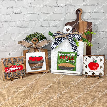 Janet's Craft Corner Apple Squares features four farmhouse-style wood signs with apple designs, displayed on burlap before a white brick wall—ideal DIY home decor accents.
