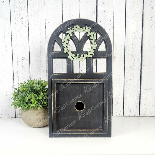The Arch Window Interchangeable Frame for Squares by Janet's Craft Corner, featuring a rustic black finish and green wreath accent, sits by a potted plant—ideal DIY farmhouse décor against white wood-panel backgrounds.