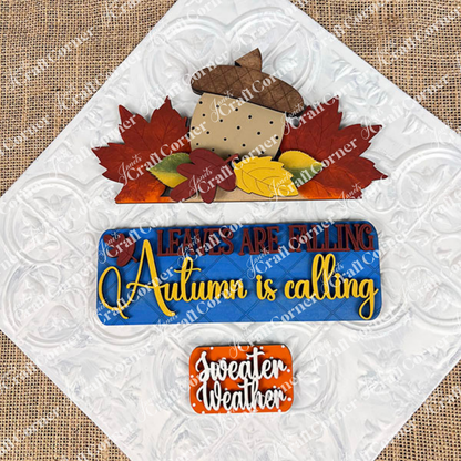 Autumn-themed decorative signs from Janet's Craft Corner, featuring the DIY home decor kit "Autumn Is Calling Interchangeable Insert." The top sign showcases an acorn with leaves and the phrase "Leaves are falling," while the customizable middle sign states, "Autumn is calling," providing an interchangeable touch. The bottom sign reads, "Sweater weather," embellished with leaves.