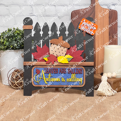 Janet's Craft Corner offers the "Autumn Is Calling Interchangeable Insert," a DIY home decor kit on a black garden gate, featuring with vibrant leaves and an acorn. The sign displays the message "LEAVES ARE FALLING, Autumn is Calling, sweater weather" and includes an interchangeable insert. It is beautifully set against a brick wall with wooden boards and a candle in the background.