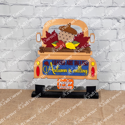 Janet's Craft Corner offers the "Autumn Is Calling Interchangeable Insert," a DIY home decor kit, featuring a decorative piece styled like the back of a copper truck with vibrant leaves and an acorn. The sign displays the message "LEAVES ARE FALLING, Autumn is Calling, sweater weather" and includes an interchangeable insert. It is beautifully set against a brick wall with wooden boards and a candle in the background.