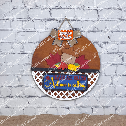 Janet's Craft Corner offers the "Autumn Is Calling Interchangeable Insert," a DIY home decor kit on a door hanger, featuring with vibrant leaves and an acorn. The sign displays the message "LEAVES ARE FALLING, Autumn is Calling, sweater weather" and includes an interchangeable insert. It is beautifully set against a brick wall with wooden boards and a candle in the background.