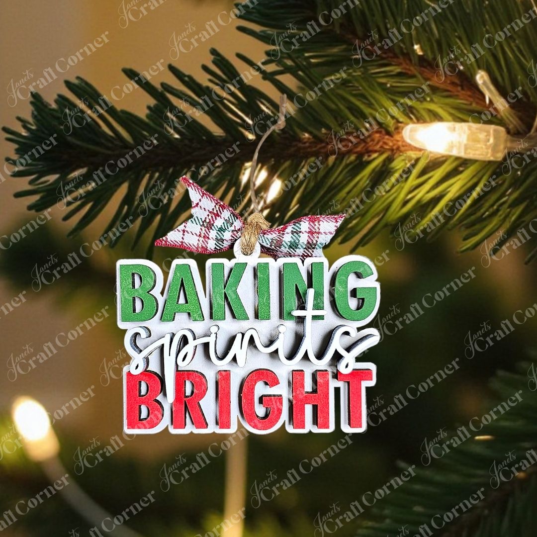 This individually painted ornament, named "Baking Spirits Bright" from Janet's Craft Corner, features the cheerful phrase in green, white, and red. Adorned with a plaid ribbon, it is beautifully displayed amidst evergreen branches and pinecones on a rustic wooden surface.