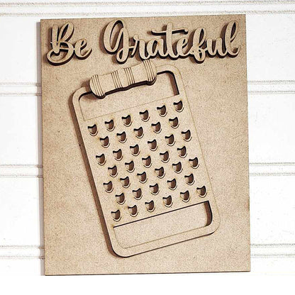 This delightful home decor piece, part of the "Be Grateful Kitchen Interchangeable Sign" by Janet's Craft Corner, showcases a "Be Grateful" message above a cheese grater design. The light wooden art, with its detailed cut-out features, is elegantly displayed on a white shiplap background, making it a perfect addition to any DIY home decor craft kit collection.