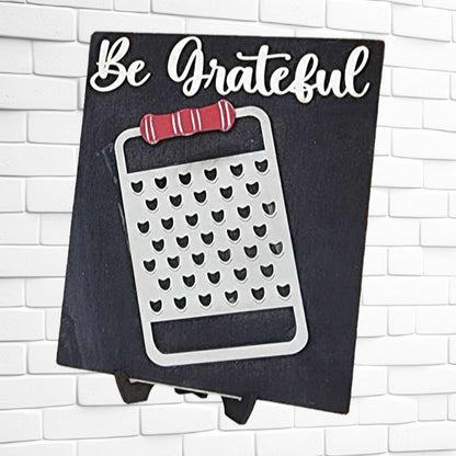 The "Be Grateful Kitchen Interchangeable Sign" DIY home decor craft kit from Janet's Craft Corner showcases a chalkboard sign with "Be Grateful" elegantly scripted at the top, complemented by a metallic cheese grater featuring a red handle beneath. This charming piece is set against a white brick wall, making it perfect for adding personality to your space or incorporating into your DIY craft projects.