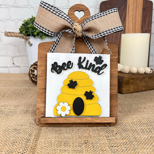 The Bee Swappable Squares by Janet's Craft Corner is a handmade decorative sign with a yellow beehive, bees, white flower, and "Bee Kind" text, finished with a gingham bow on a wooden stand for charming home decor.