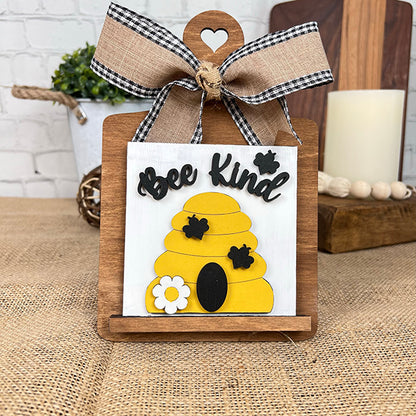 The Bee Swappable Squares by Janet's Craft Corner is a handmade decorative sign with a yellow beehive, bees, white flower, and "Bee Kind" text, finished with a gingham bow on a wooden stand for charming home decor.