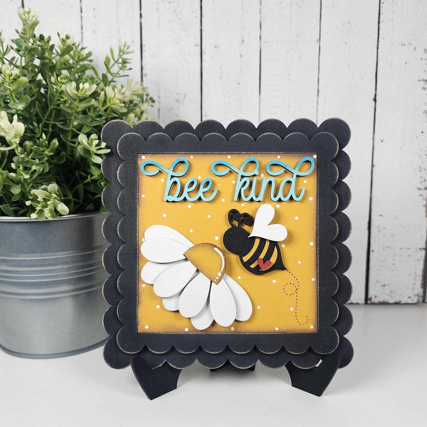 Janet's Craft Corner’s Bee Swappable Squares sign features "bee kind" in cursive, a handmade 3D white flower, and a yellow-black bee on a yellow background—displayed on an easel, perfect for home decor.