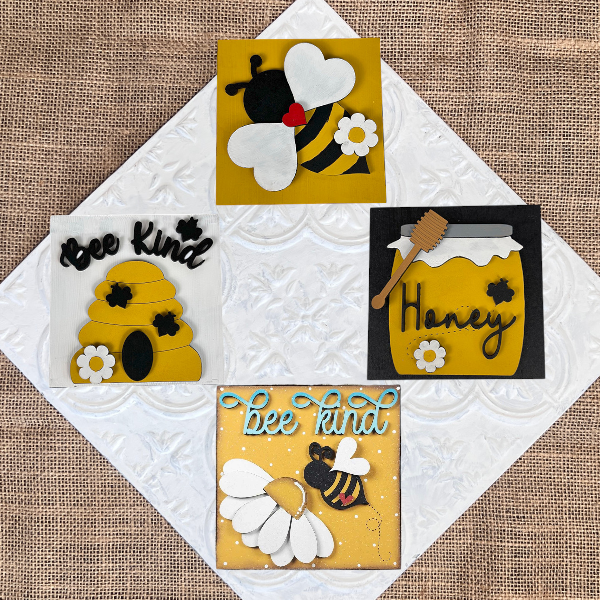 The Bee Swappable Squares DIY kit by Janet's Craft Corner includes four burlap-backed designs—a bee, a honey jar labeled "Honey," a beehive, and "bee kind" text—arranged on a textured white tile for charming home decor.