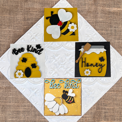 The Bee Swappable Squares DIY kit by Janet's Craft Corner includes four burlap-backed designs—a bee, a honey jar labeled "Honey," a beehive, and "bee kind" text—arranged on a textured white tile for charming home decor.