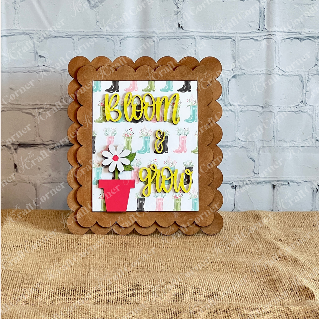 Janet's Craft Corner Spring Switchable Farmhouse Signs DIY Kit features a scalloped wooden frame with a card showing colorful rain boots, a red flowerpot with a white flower, and yellow "Bloom & grow" text on a white brick wall background.