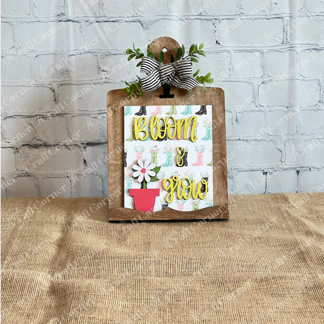 Janet's Craft Corner Spring Switchable Farmhouse Signs DIY Kit includes a wooden clipboard with a bow and greenery, featuring a "Bloom & Grow" sign with a pink potted flower—perfect for tiered tray styling on burlap and white brick.