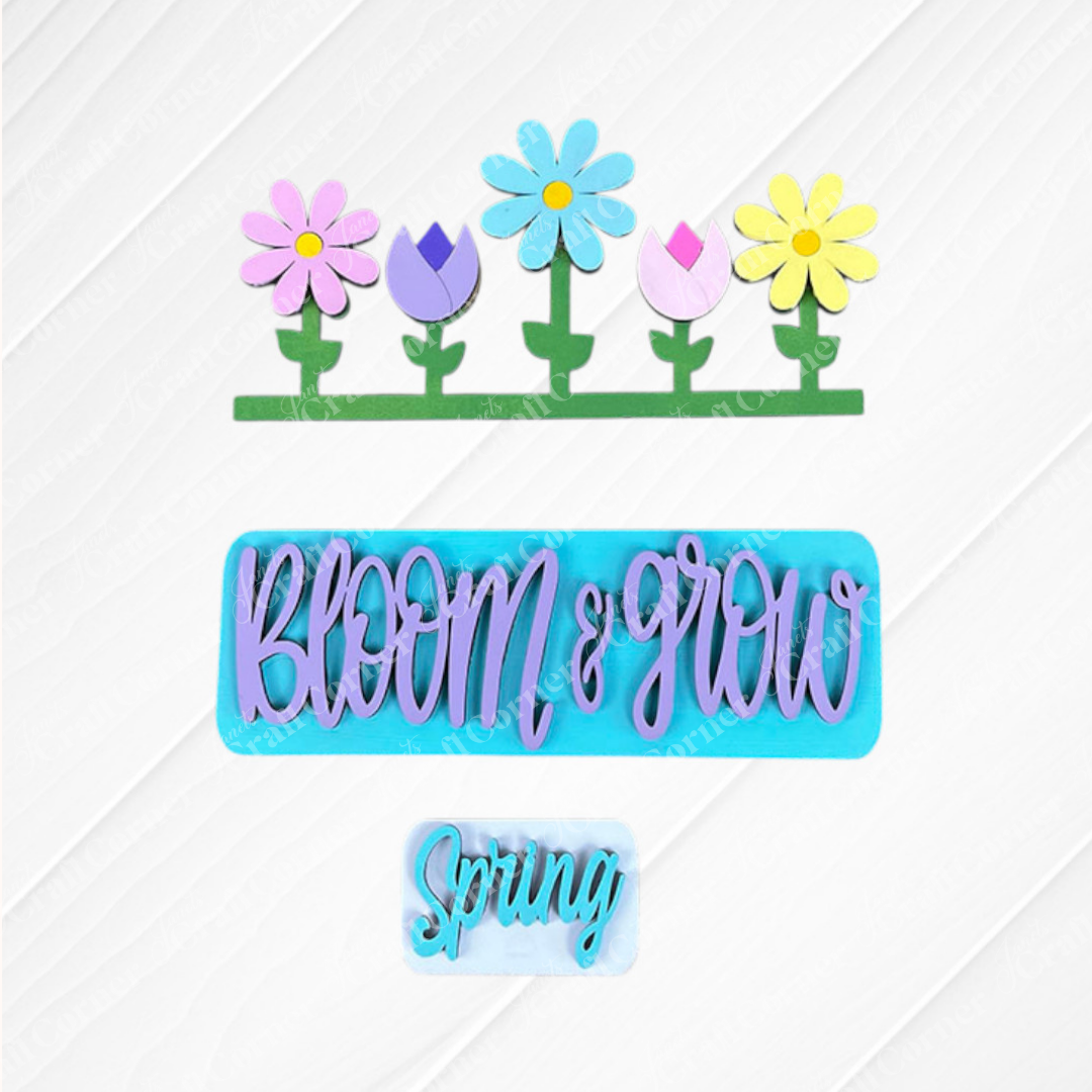 The Spring Blooms Interchangeable Insert from Janet's Craft Corner features colorful wooden cutouts on a white wood background. The top showcases pastel flowers, the middle highlights "Bloom & Grow" in purple on a teal background, and the bottom displays "Spring" in white against teal. It's perfect for home décor or as a hand-painted DIY craft kit.