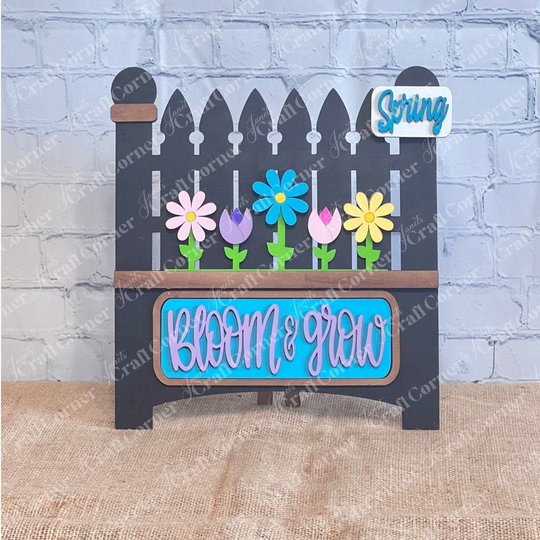 The "Bloom & Grow" Interchangeable Insert by Janet's Craft Corner is a DIY seasonal home decor craft kit featuring a wooden picket fence sign with colorful flowers and a small "Spring Blooms" message, set against a brick wall with burlap for rustic charm.