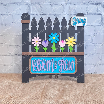 The "Bloom & Grow" Interchangeable Insert by Janet's Craft Corner is a DIY seasonal home decor craft kit featuring a wooden picket fence sign with colorful flowers and a small "Spring Blooms" message, set against a brick wall with burlap for rustic charm.