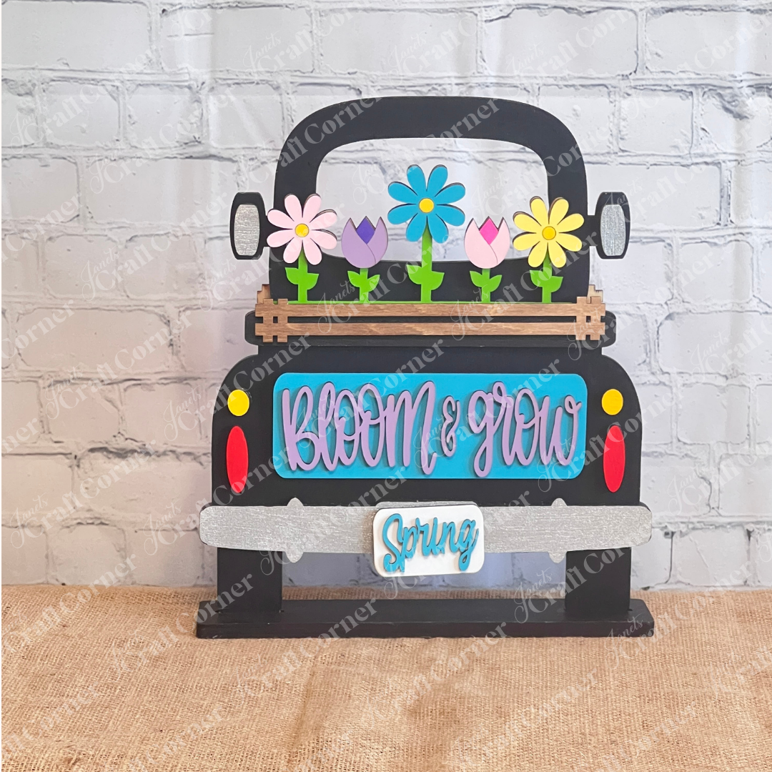 This antique wooden truck display from Janet's Craft Corner features a blue area with "Bloom & Grow" in pink, colorful Spring Blooms in the bed, "Spring" on the bumper, all set on burlap against a brick-patterned backdrop as part of the Bloom & Grow Interchangeable Insert DIY kit.