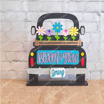 This antique wooden truck display from Janet's Craft Corner features a blue area with "Bloom & Grow" in pink, colorful Spring Blooms in the bed, "Spring" on the bumper, all set on burlap against a brick-patterned backdrop as part of the Bloom & Grow Interchangeable Insert DIY kit.
