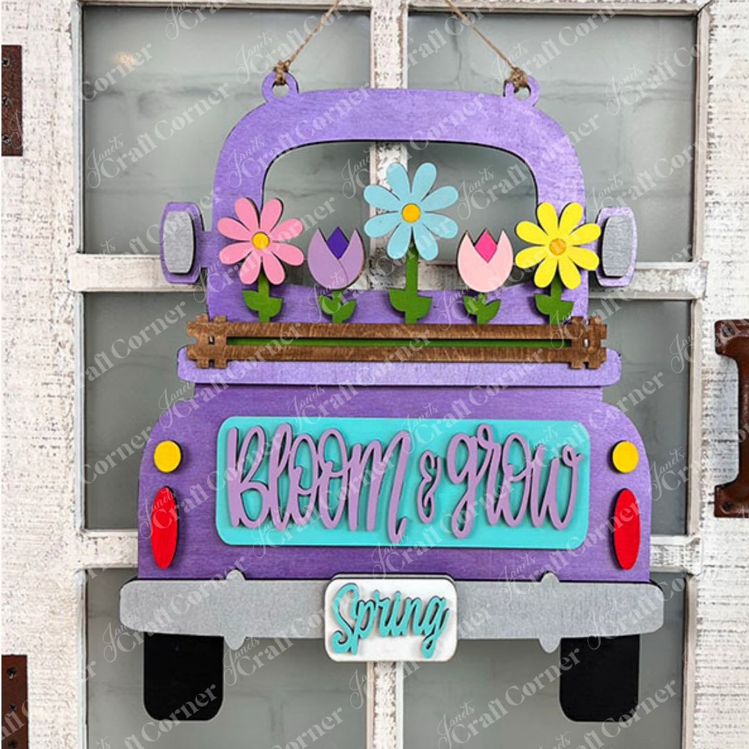 Bring charm to your home décor with Janet's Craft Corner's Spring Blooms Interchangeable Insert. This delightful DIY home decor kit features a hand-painted purple truck brimming with vibrant flowers and a whimsical "Bloom & grow" message. A quaint little "Spring" sign dangles from the bumper, all beautifully set against a rustic white door backdrop.