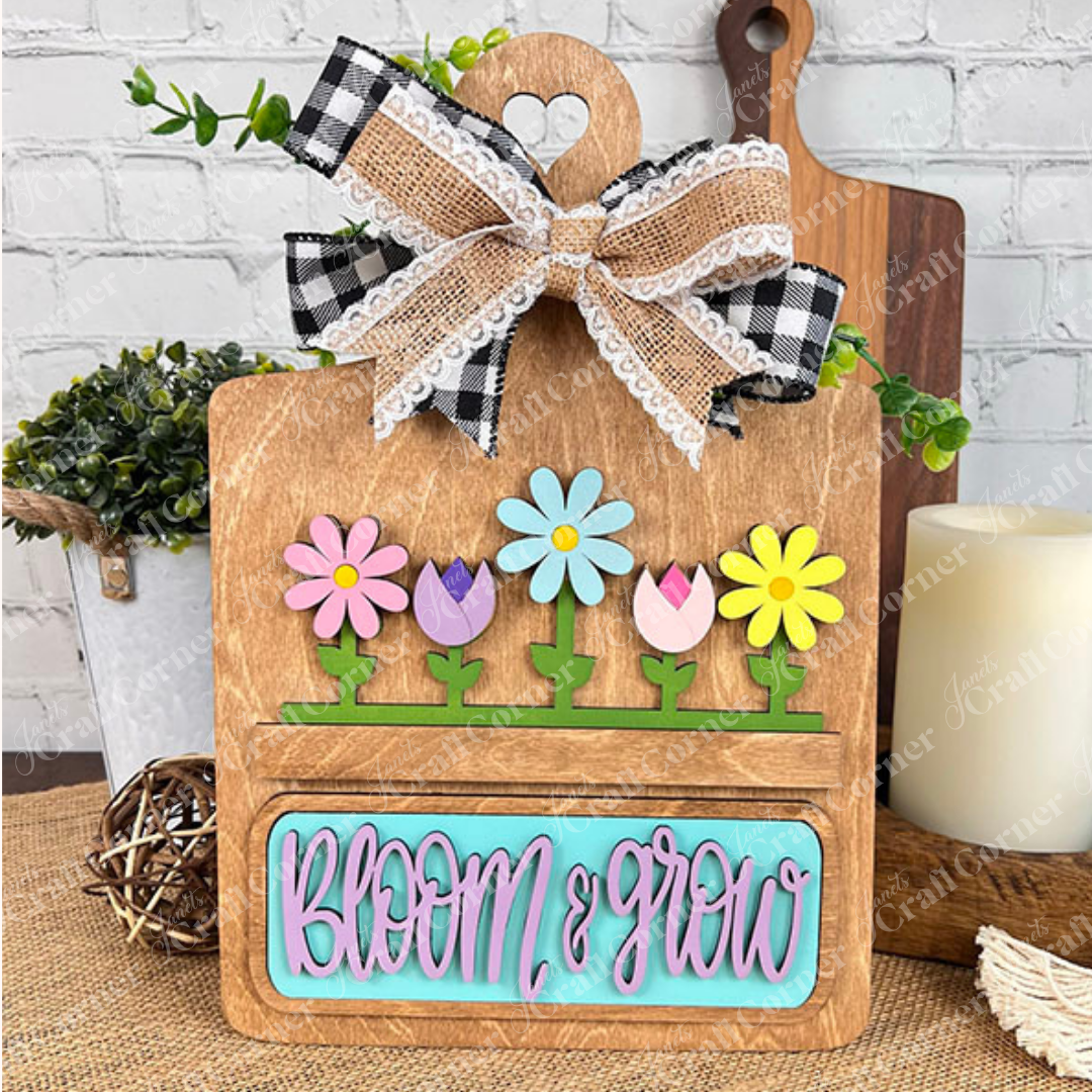 Introducing the Spring Blooms Interchangeable Insert DIY home decor kit from Janet's Craft Corner. This hand-painted craft set includes a wooden cutting board display adorned with a charming checkered lace bow and features vibrant flower cutouts. The rustic backdrop beautifully showcases the phrase "Bloom & Grow" in blue. It's perfect for enhancing your home décor and comes with candle, basket, and wood accents to complete the display.